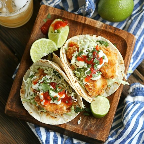 Crispy, golden-brown fish tacos topped with shredded cabbage, creamy white sauce, fresh cilantro, and spicy salsa, served on lightly toasted corn tortillas with lime wedges on a rustic wooden plate. A refreshing cold drink and striped napkin complete this vibrant Baja-style meal.